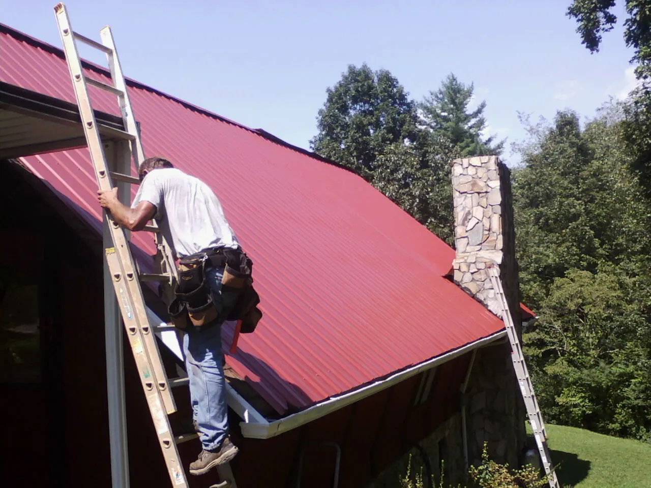 Roofer on a ladder beside a red metal roof and stone chimney — Soffit & Fascia Repair by Roofing Brothers in State College